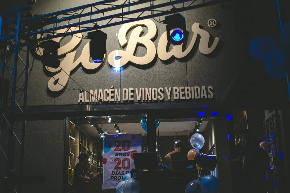 La misma fachada esa noche, con el cartel del local iluminado por las cabezas móviles con haz azul y gente adentro durante la inauguración